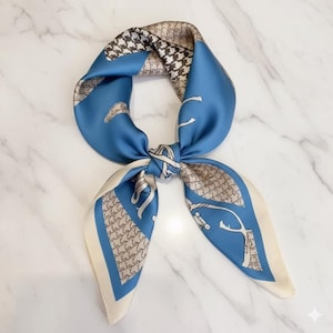 Peut inclure: Un foulard carré en soie avec un motif bleu, crème et marron. Il présente un motif pied-de-poule, des illustrations équestres et un design noué. Le foulard est plié et noué, mettant en valeur ses différents motifs et couleurs.