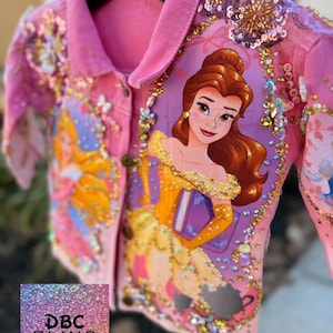 Op de afbeelding: Roze spijkerjasje met een cartoonillustratie van Belle uit Belle en het Beest, versierd met pailletten en strass. De jas heeft een knoopsluiting en de tekst "DBC BLING".