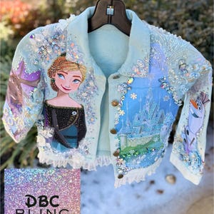 Puede incluir: Chaqueta vaquera azul claro con un diseño pintado a mano de una princesa y un castillo. La chaqueta está adornada con pedrería y abalorios. El texto "DBC BLING" es visible en un cuadrado brillante.
