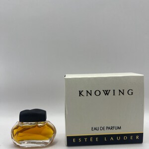May include: A miniature perfume bottle with a black cap and amber liquid is positioned beside a white box. The box is labelled "KNOWING" with "EAU DE PARFUM" and "ESTEE LAUDER" printed below. The perfume bottle is a small, clear glass container.