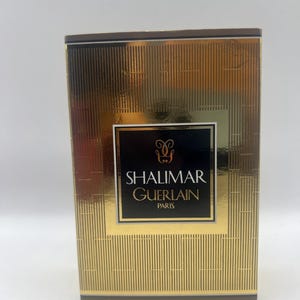 Könnte beinhalten: Eine goldfarbene Parfümbox mit vertikalen und horizontalen strukturierten Linien. Die Box hat ein schwarzes Etikett mit den Worten "SHALIMAR GUERLAIN PARIS" in weißer Schrift. Die Oberseite und die Kanten der Box sind dunkelbraun.