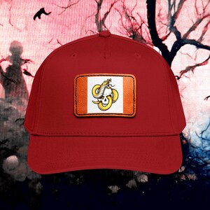 Könnte beinhalten: Eine rote Baseballkappe mit einem rechteckigen Aufnäher auf der Vorderseite. Der Aufnäher zeigt einen weißen Hintergrund mit einer gelben Schlange, die sich um einen weißen Totenkopf windet. Der Aufnäher hat einen braunen Rand.