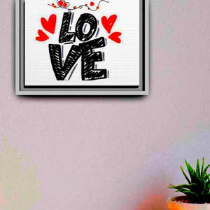 以下が含まれることがあります： 「LOVE」の文字が大きく黒い手書き文字で描かれた、額装されたキャンバスアート。赤いハートと装飾的な線が白い背景を飾っています。フレームはシルバーで、ピンク色の壁に飾られています。
