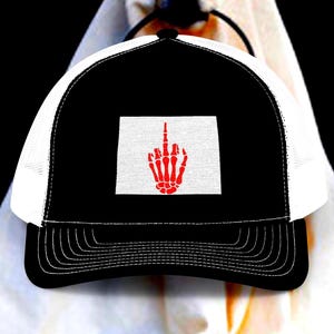 May include: A black and white trucker hat featuring a rectangular patch on the front panel. The patch displays a red graphic of a skeletal hand with the middle finger extended in a gesture. The hat has a solid black brim and a white mesh back.