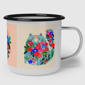 K&ouml;nnte beinhalten: Ein wei&szlig;er Emaillebecher mit schwarzem Rand und silbernem Henkel. Der Becher zeigt eine grafische Illustration einer hellblauen Katze, die mit leuchtend roten, rosa und blauen Blumen bedeckt ist, neben einem Blumenarrangement auf pfirsichfarbenem Grund.