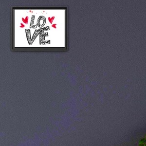以下が含まれることがあります： 黒の手書き文字で「LOVE」と書かれた額入りのプリント。赤いハートと飛沫が単語の上を飾っています。フレームは黒で、背景は白です。