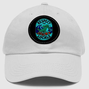 Puede incluir: Una gorra de béisbol blanca con un parche circular negro en la parte delantera. El parche muestra una foca verde sobre rocas rojas frente a un océano azul con el texto RESPECT THE LOCALS en letras azules alrededor del gráfico.