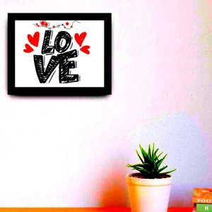 以下が含まれることがあります： 黒いスケッチフォントで「LOVE」という言葉が書かれた額入りのアートプリント。赤いハートのアクセントと黒いフレームが白い背景を囲んでいます。アートワークは白い壁に飾られており、下に小さな鉢植えと本があります。