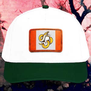 Puede incluir: Una gorra de béisbol blanca con visera verde oscuro y un parche rectangular en la parte delantera. El parche presenta una ilustración gráfica de una serpiente amarilla enroscada alrededor de un cráneo humano blanco sobre un fondo gris claro con bordes naranjas.