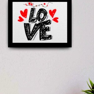 以下が含まれることがあります： 手描きのスタイルで「LOVE」という言葉が描かれた黒い額縁のアートワーク。 黒い文字は赤いハートと装飾的な要素で縁取られています。 アートワークは白い背景にあります。