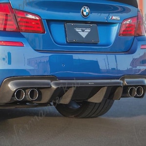 Op de afbeelding: Achteraanzicht van een blauwe BMW M5 met een zwarte diffuser en dubbele uitlaatpijpen. De auto heeft een Vorsteiner kentekenplaat en rode achterlichten. De foto is buiten genomen.