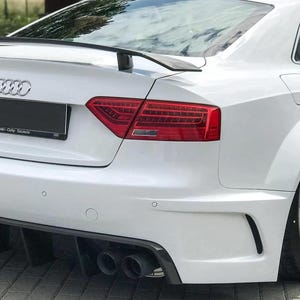 Könnte beinhalten: Weißes Audi S5 Coupé mit schwarzem Heckspoiler und Diffusor. Das Auto hat rote Rückleuchten, Doppelendrohre und ein schwarzes Nummernschild. Die Räder sind silber.
