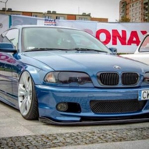 Könnte beinhalten: Ein blaues BMW-Auto mit schwarzem Kühlergrill und Nummernschild. Das Auto steht auf einer gepflasterten Fläche. Das Auto hat ein tiefergelegtes Fahrwerk und Sonderräder. Der Hintergrund umfasst ein weißes Auto und ein Banner mit rotem Text.