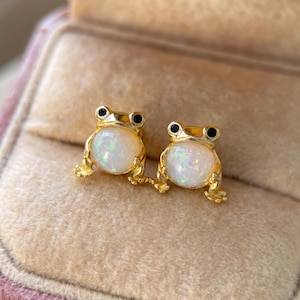May include: A pair of gold-toned stud earrings shaped like frogs. Each frog features a round, iridescent white opal-like stone as its body, with small black accents for the eyes. The metal has a polished finish, highlighting the frog's limbs.