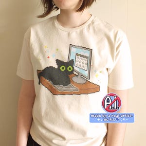 May include: A cream-colored t-shirt featuring a pixel art graphic of a black cat sitting on a desk in front of a computer monitor. The screen displays the text AAAA AAAA AAAA. A circular logo with the text NO A.I. and a blue banner with the text made by a real artist- no A.I. are visible.