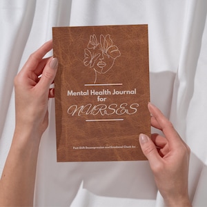 May include: A brown journal with the text "Mental Health Journal for NURSES" in white script. The cover features a line drawing of a face with butterfly accents. The journal is held by two hands against a white fabric background.