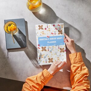 May include: A "Monthly Nurse Shift Planner" with a light blue banner, held by a person wearing an orange shirt. The planner has a white background with medical-themed illustrations. A gray notebook and drinks are also visible.