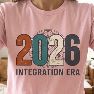 May include: A light pink t-shirt with the text "2026 INTEGRATION ERA" in a retro design. The numbers are in orange, teal, white, and gray. The shirt is a crew neck and has short sleeves.