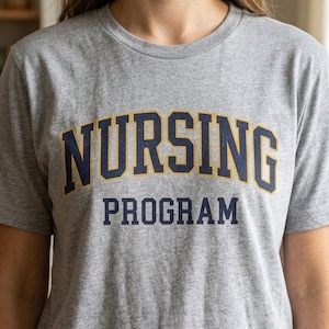 Peut inclure: T-shirt gris chiné avec l'inscription "NURSING PROGRAM" en bleu marine et or. Le t-shirt a un col rond et des manches courtes. L'image montre le devant du vêtement.