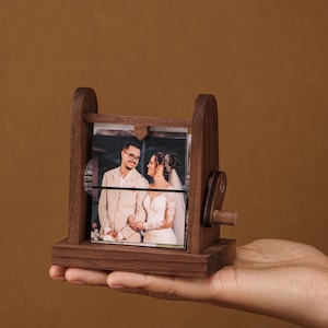 May include: A wooden photo display with a rotating mechanism, displaying wedding photos. The dark brown frame holds multiple images, with a crank on the side for turning. The photos feature a couple in wedding attire.