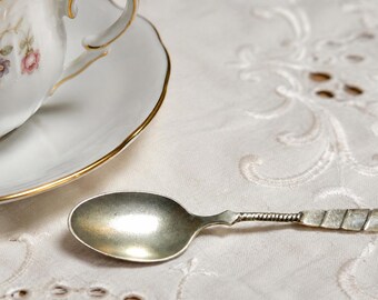 May include: A white porcelain teacup and saucer set featuring a delicate pink rose and green leaf floral pattern with gold trim, accompanied by a small silver-toned metal spoon with a textured, spiraled handle design.