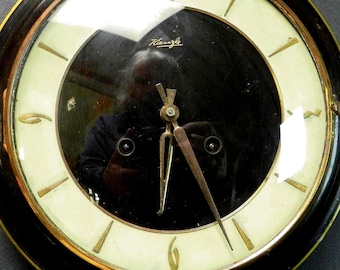 May include: A round wall clock featuring a black center dial with the Kienzle brand name in gold, surrounded by a cream-colored ring with gold-toned hour markers and numerals. The clock has a dark frame, gold hands, and a gold-toned top mount.