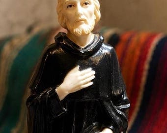 May include: A figurine of a bearded figure wearing a black robe with one leg exposed, standing on a rectangular base with the text St. Peregrine inscribed on the front. The figure has light-colored skin and hair, with one hand placed on the chest.