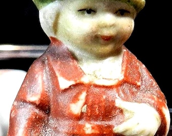 May include: A small, vintage ceramic figurine of a person wearing a red dress and a green hat. The figurine is holding a green pouch. The figure has a pale complexion and is standing upright.