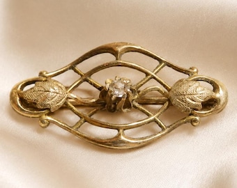 May include: An oval-shaped gold-tone brooch featuring an openwork lattice design. The piece is accented with two textured leaf motifs on the left and right sides and a single clear, faceted stone set in the center of the metal framework.