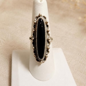 May include: A silver ring featuring a long, narrow, oval-shaped black stone set vertically. The metal setting is decorated with a series of small silver spheres and a twisted rope-like border surrounding the central black gemstone.