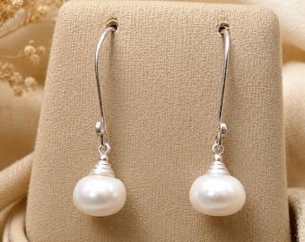 May include: A pair of drop earrings featuring smooth, white, oval-shaped pearls suspended from curved silver-toned metal hooks. Each pearl is capped with a small, ribbed silver-toned metal accent where it connects to the long, slender wire hook.