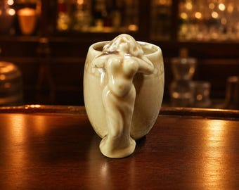May include: A beige ceramic mug featuring a sculpted figure of a person with arms raised above their head, serving as the handle. The mug has a glossy, crackled glaze finish and a circular pattern embossed around the upper rim of the vessel.