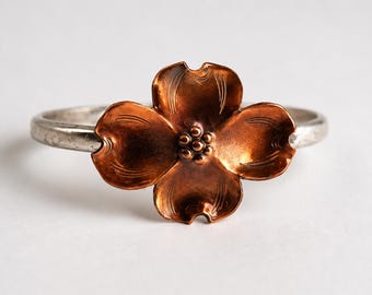 May include: A silver ring featuring a copper-colored flower design. The flower has four petals and a textured center. The ring is set against a white background.