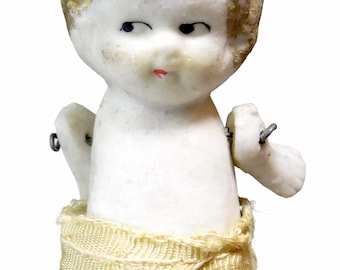May include: A vintage bisque doll with a painted face, black eyes, and a red mouth. The doll has a yellow fabric wrap around its waist and jointed arms. The doll's hair is painted gold and green. The doll is on a white background.