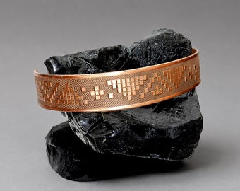 May include: A copper cuff bracelet featuring a repeating geometric pattern of small squares and triangles etched into the metal surface. The band has a smooth, polished finish and a consistent width, showcasing a metallic orange-brown color.