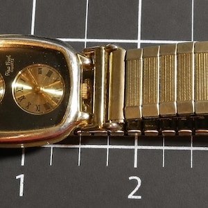 May include: A gold-tone wristwatch with a rectangular case and a black dial featuring two circular sub-dials, one with Arabic numerals and one with Roman numerals. The watch has an expandable link bracelet and measures approximately 3.5 inches long.
