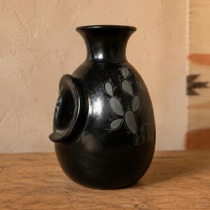 May include: A black ceramic vase with a smooth, polished finish and a rounded body. The vase features a prominent circular side handle and an etched design of a prickly pear cactus on its surface, showcasing fine, light-colored line details.