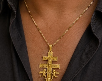 May include: A gold-toned metal pendant necklace featuring a multi-barred cross design. The pendant displays a central figure of Jesus on the cross, the text CARA VACA, two winged figures, and a skull at the base, suspended from a thin gold chain.