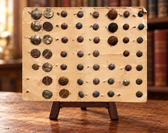 May include: A rectangular, aged, off-white cardboard display card featuring a grid of numerous small, round, metallic buttons in various shades of brown, bronze, and dark grey, supported by a small, dark brown wooden easel stand.