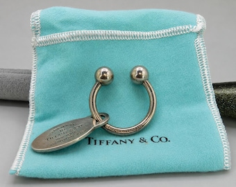 May include: A sterling silver Tiffany & Co. horseshoe-shaped keychain with two spherical ends and an oval tag engraved with Please Return To Tiffany & Co. New York 925 Sterling Silver, resting on a light blue fabric pouch with the brand name.
