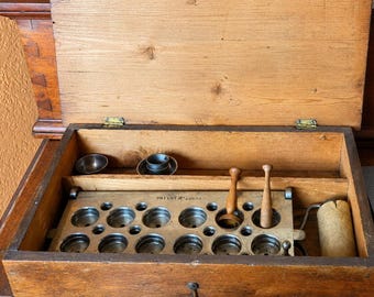May include: Open wooden box with a hinged lid, containing various metal and wooden tools. The box has a metal tray with circular openings, two wooden tools, and small metal cups. The box is made of light brown wood.