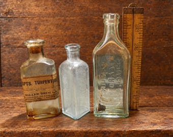 May include: Three clear glass bottles of varying heights and shapes. The left bottle has a tan paper label reading SPTS. TURPENTINE MILLER DRUG CO. PHILADELPHIA, PA. LANCASTER, PA. WILMINGTON, DEL. TRENTON, N.J. A wooden folding ruler stands beside them.