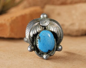 May include: A silver ring featuring a central oval-shaped blue turquoise stone. The metal setting is decorated with embossed leaf patterns and small spherical accents at the top and bottom, creating a detailed metallic frame around the stone.