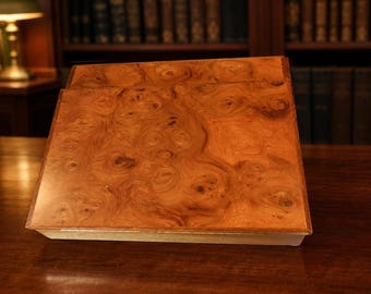 May include: A slanted wooden book stand featuring a polished burl wood grain surface with swirling brown and amber patterns. The rectangular stand has a smooth, glossy finish and a flat base, designed to hold reading materials at an angle.