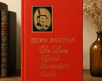 May include: A red hardcover book titled "OLD MR. BOSTON De Luxe Official Bartender's Guide." The cover features a gold-framed portrait of a man in a top hat. The book is standing upright on a wooden surface.