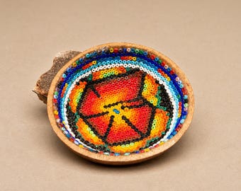 May include: A handcrafted, round bowl with a vibrant beaded design. The bowl features a central starburst pattern in red, orange, and yellow, surrounded by concentric rings of blue, white, and black beads. The bowl rests on a piece of wood.