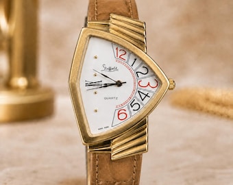 May include: A gold-tone quartz wristwatch with a triangular case and a tan leather strap. The white dial features the brand name Stafford, black hour and minute hands, and large numerals from 1 to 6 arranged along the right side in red and black.