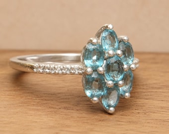 May include: A silver ring featuring a cluster of oval, light blue gemstones. The ring's band is adorned with small, clear stones. The gemstones are arranged in a floral or cluster design, creating a sparkling effect. The ring is set against a wooden surface.