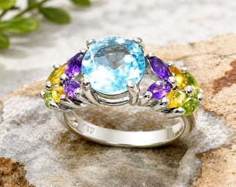 May include: A silver ring featuring a large, round, light blue gemstone as the centerpiece. The ring is adorned with smaller, colorful gemstones in shades of purple, yellow, and green. The ring is set on a textured, light brown surface.