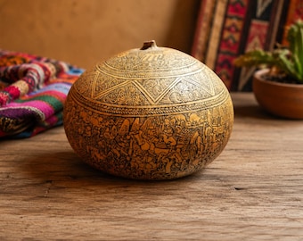 May include: A round, tan-colored gourd featuring intricate, dark-etched carvings. The surface displays detailed scenes of figures, animals, and structures, with a geometric patterned band encircling the upper portion and a small, centered stem at the top.